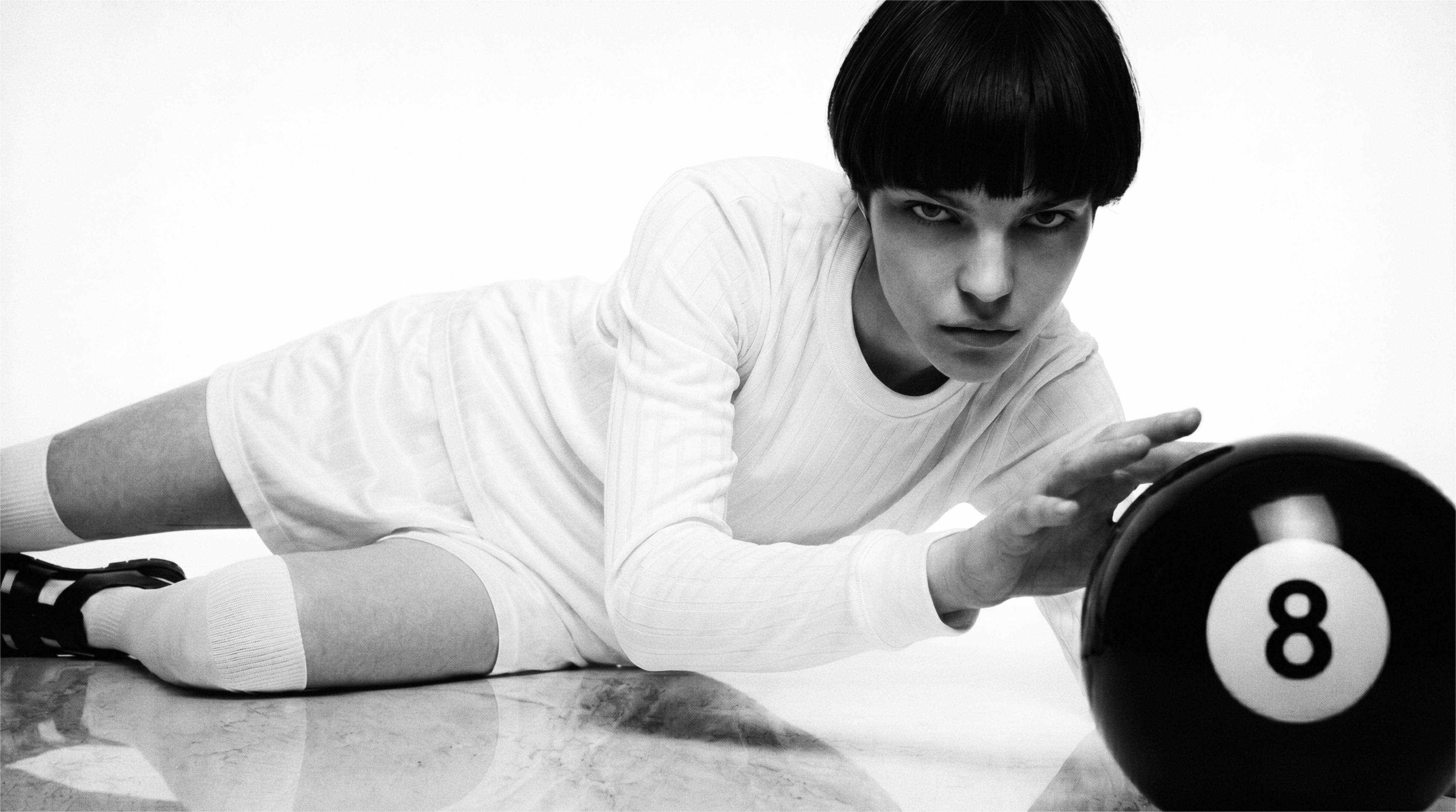 Black-and-white portrait of a model in white sportswear on a reflective studio floor, reaching toward a black 8-ball.