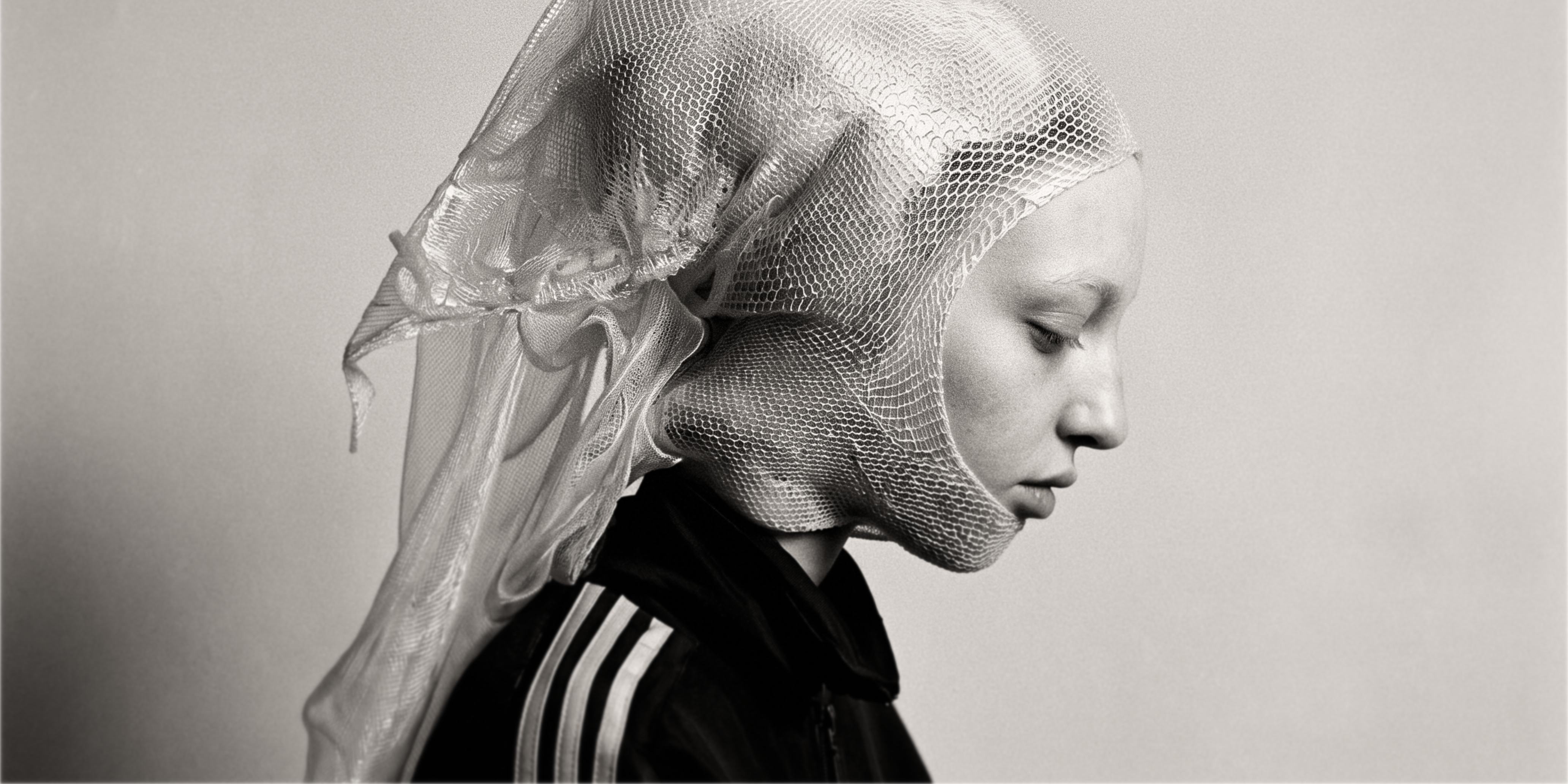 Profile portrait of a model in a black track jacket and translucent sculptural veil against a soft grey background.