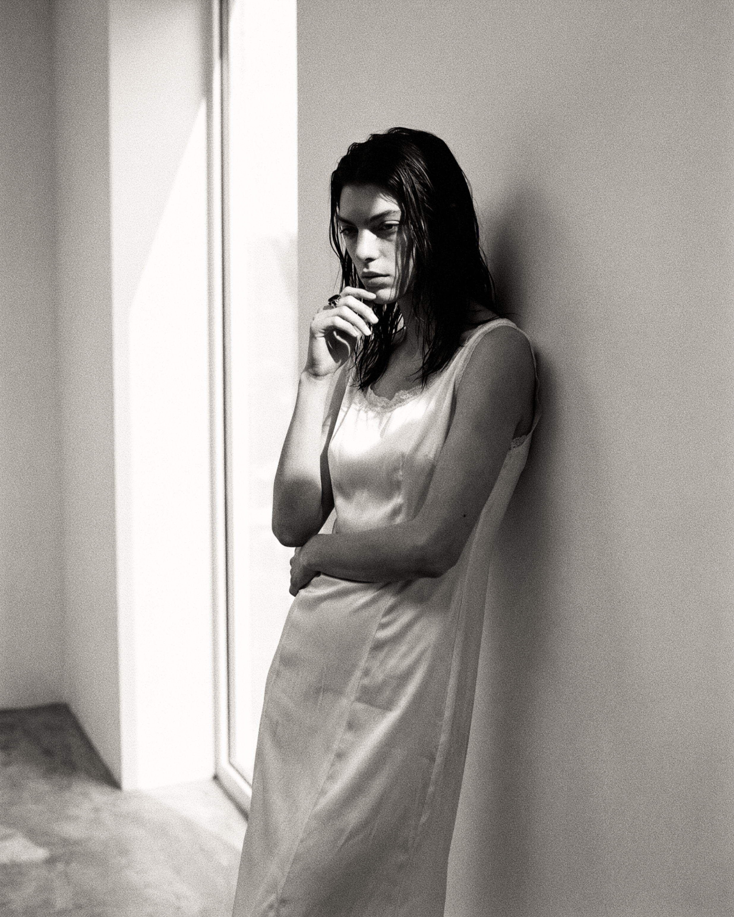A woman in a satin slip, thinking, beside a window