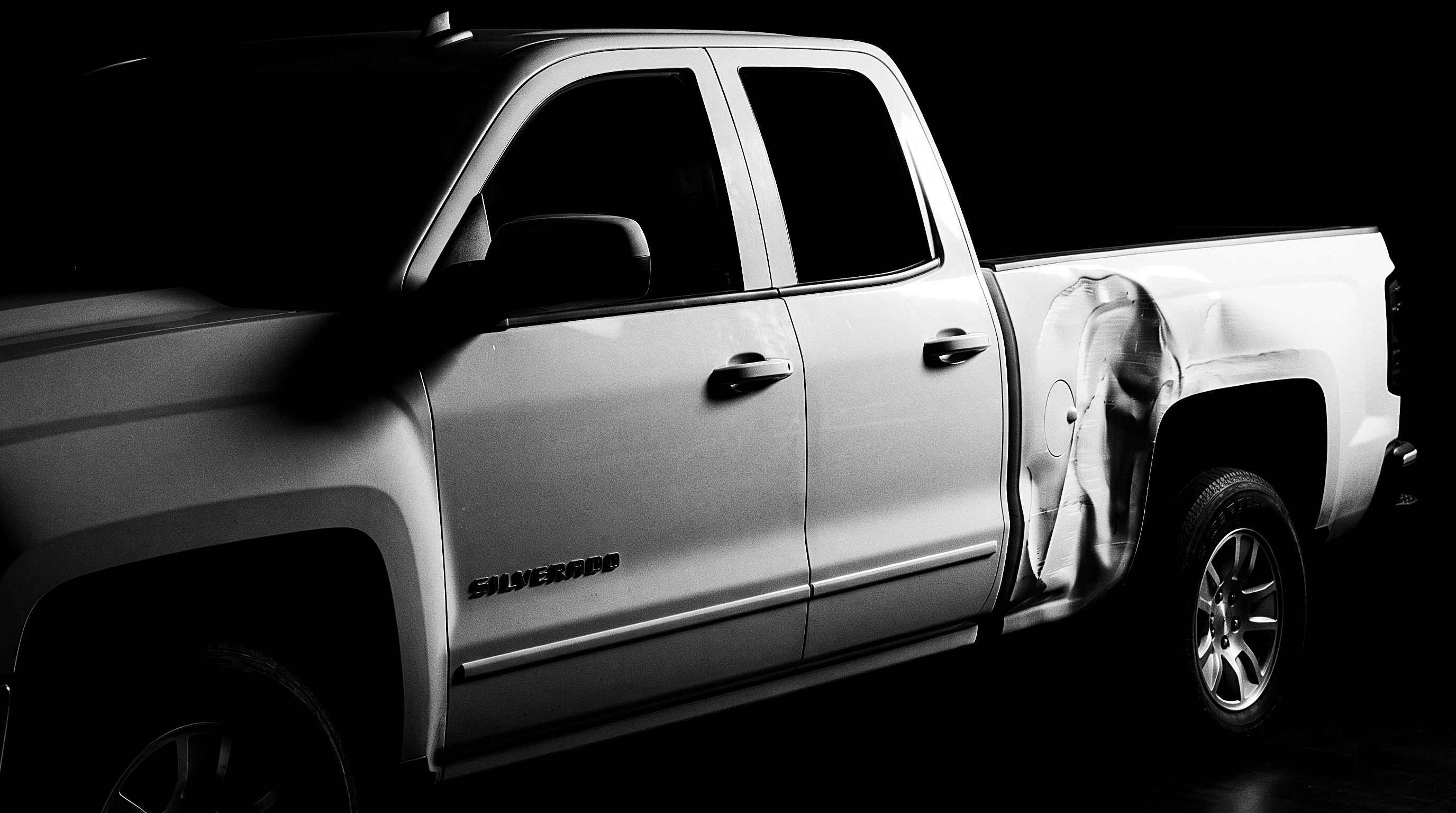 Black-and-white studio photograph of a white Chevrolet Silverado pickup against black, its rear quarter panel crumpled, the dent catching the light.