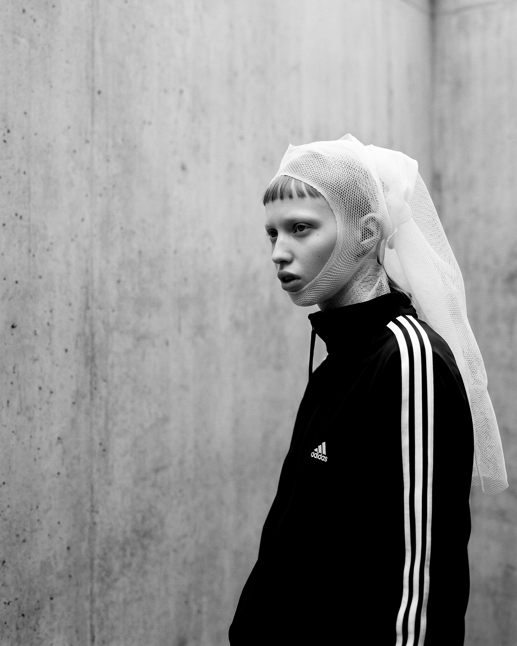 Work (Uncommissioned) — a black-and-white profile against raw concrete: a white mesh veil wrapped over the head and under the chin, worn with a black Adidas track jacket, three stripes down the sleeve