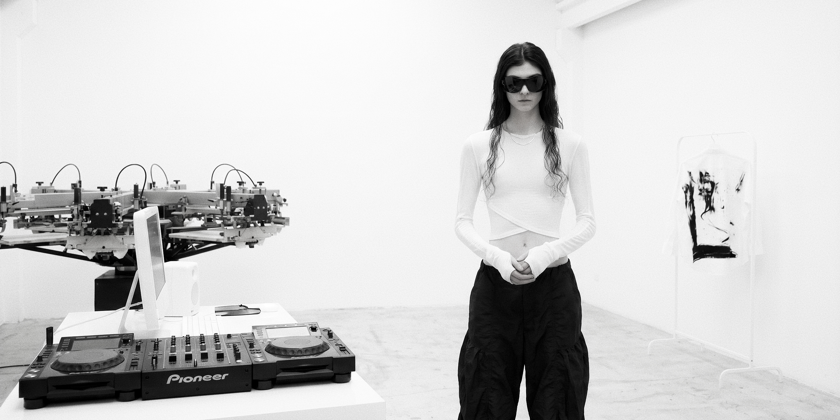 Negative Space — a white room: a screen-printing carousel at the left, Pioneer decks on a plinth, a figure in a white long-sleeve and black cargo trousers standing at the centre in wraparound sunglasses, a printed shirt hanging on a rail at the right.