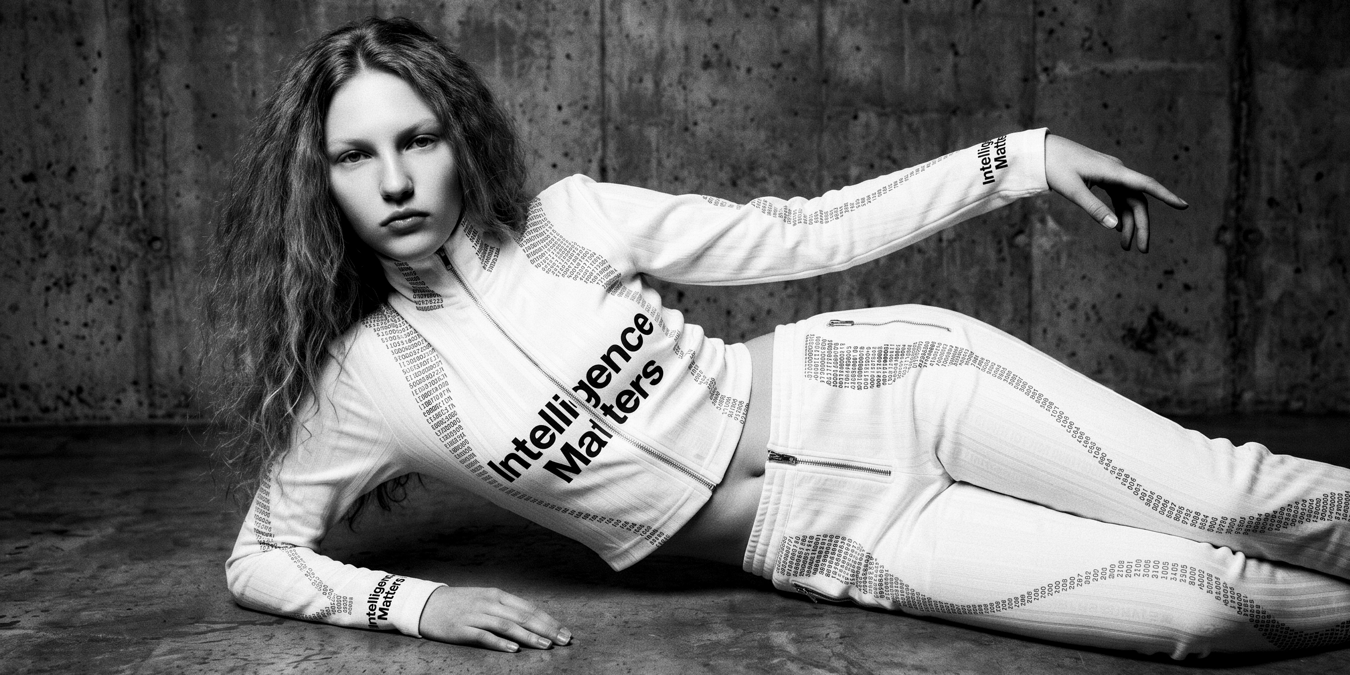 Black-and-white studio frame — a figure reclining on a concrete floor against a concrete wall, in a white zip top and trousers traced with columns of binary, Intelligence Matters set diagonally across the chest.