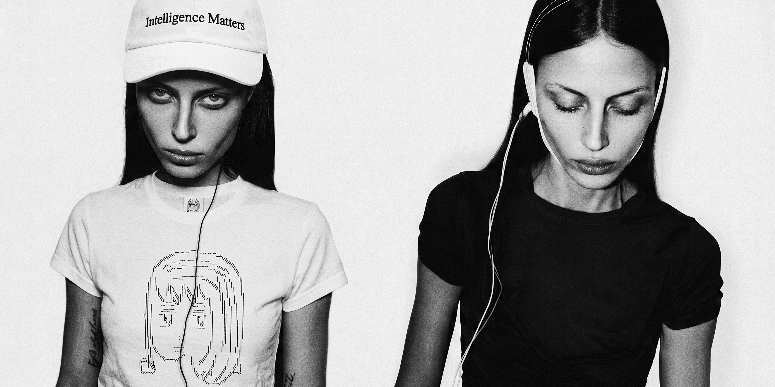 Black-and-white double portrait against white — two figures side by side: one in a white Intelligence Matters cap and a white tee printed with the studio's ASCII muse, one in black with a white wired headpiece, eyes lowered.