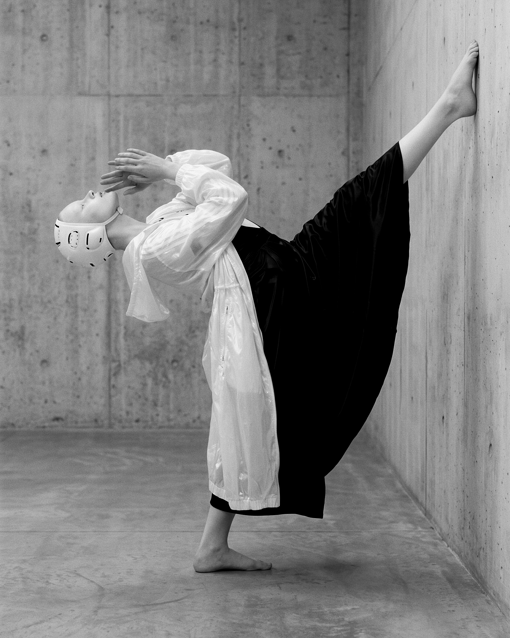 Work (Uncommissioned) — a black-and-white figure study against concrete: a dancer in a white sparring headguard, sheer white zip shell and full black skirt, one leg extended high to the wall