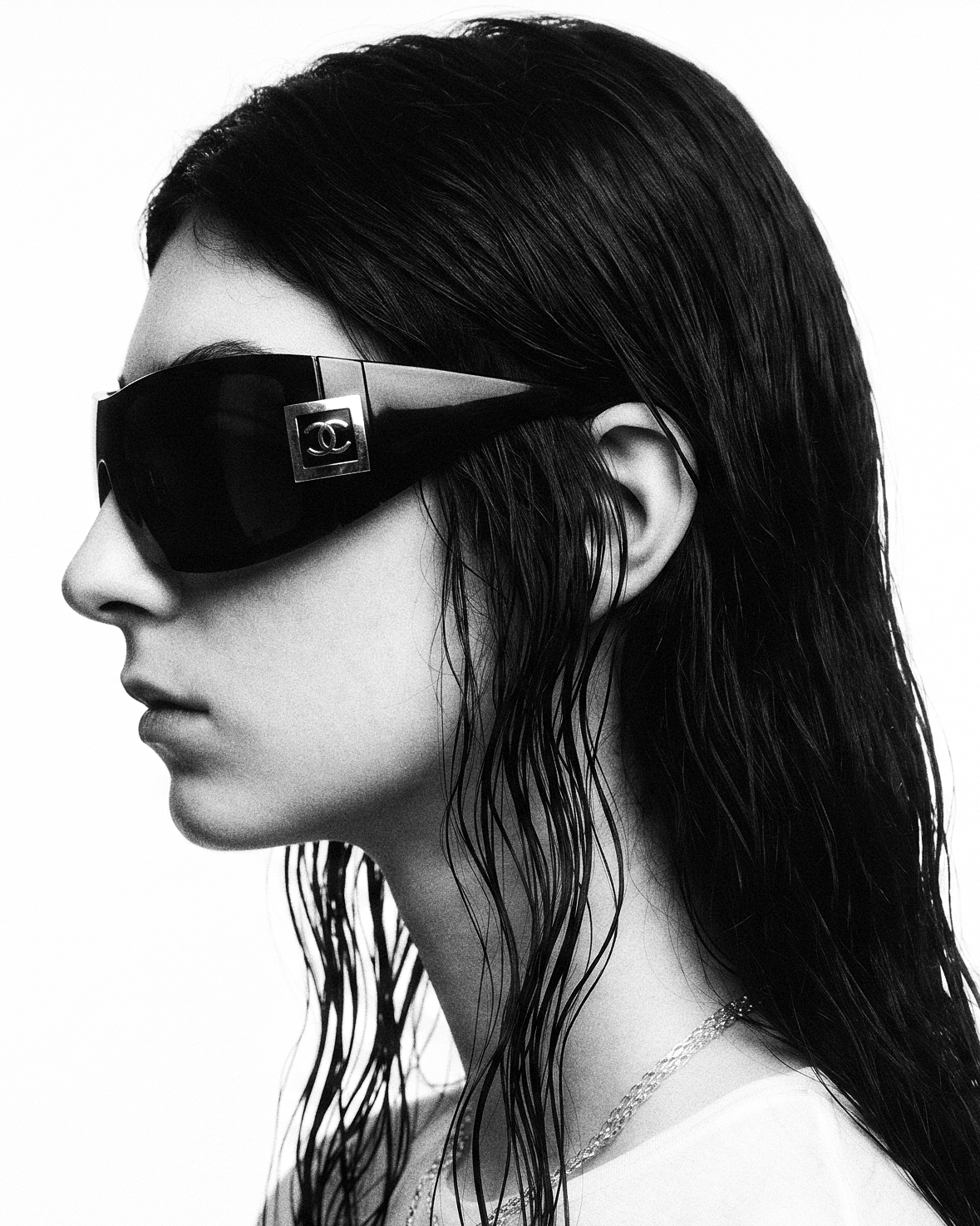 Image — a black-and-white profile against white: wet dark hair, black wraparound shield sunglasses with a silver logo plaque, a fine chain over a white tee