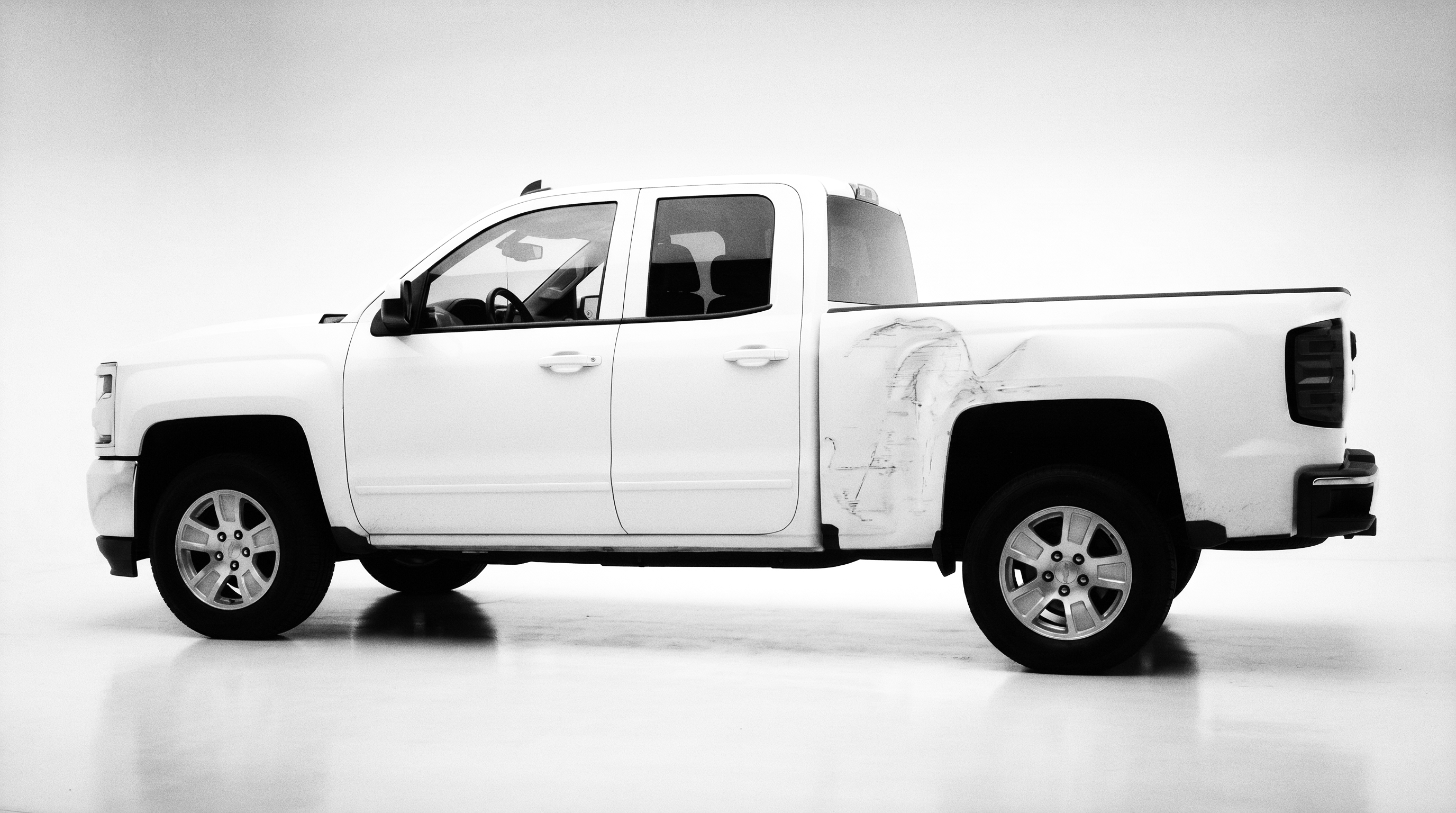 A white Chevrolet Silverado in full side profile on a white studio sweep, its flank scraped and dented behind the cab — the damage in plain daylight.