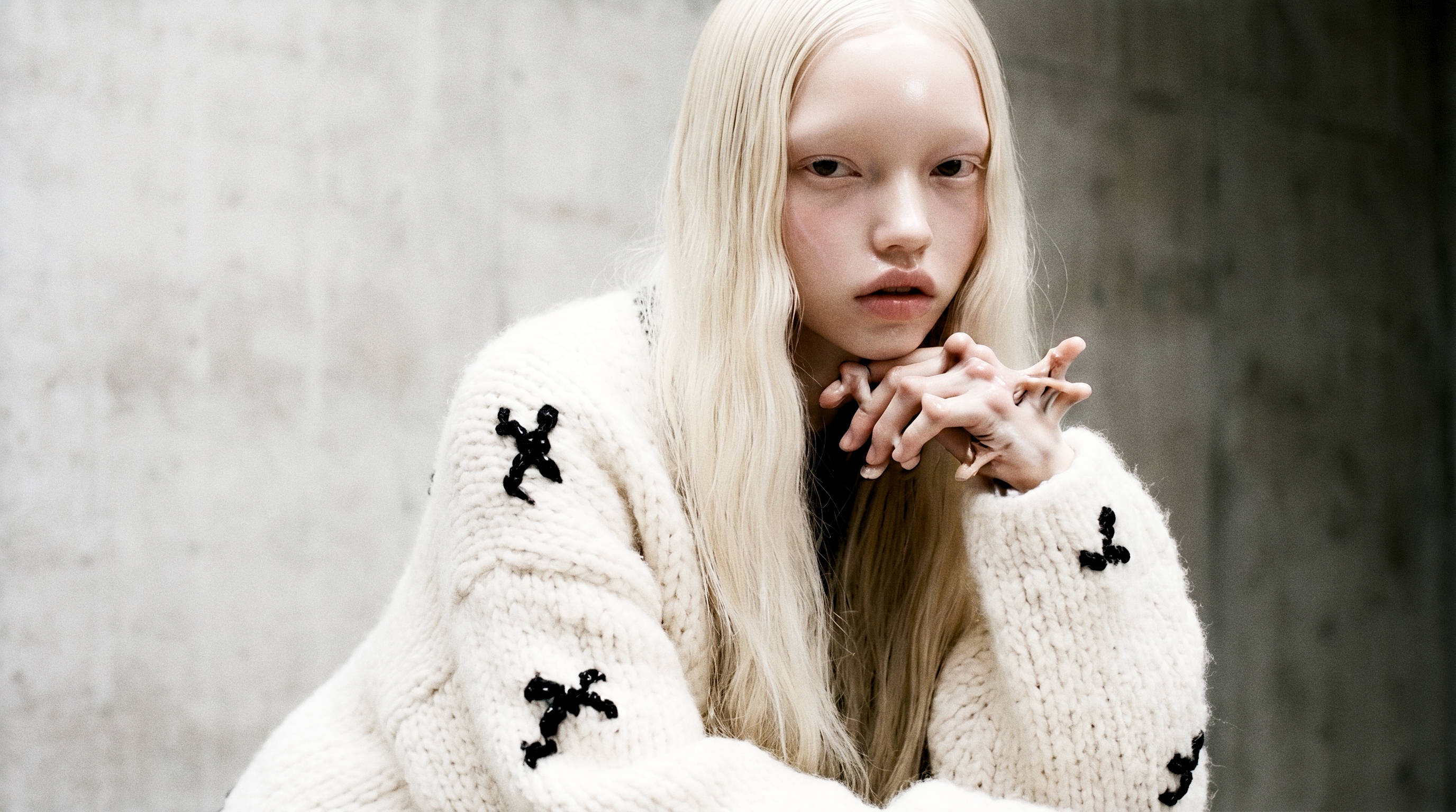 Black-and-white portrait of a pale platinum-haired figure in a chunky cream knit with black marks, chin resting on interlaced hands — the fingers tangling into one too many, the tell left in.