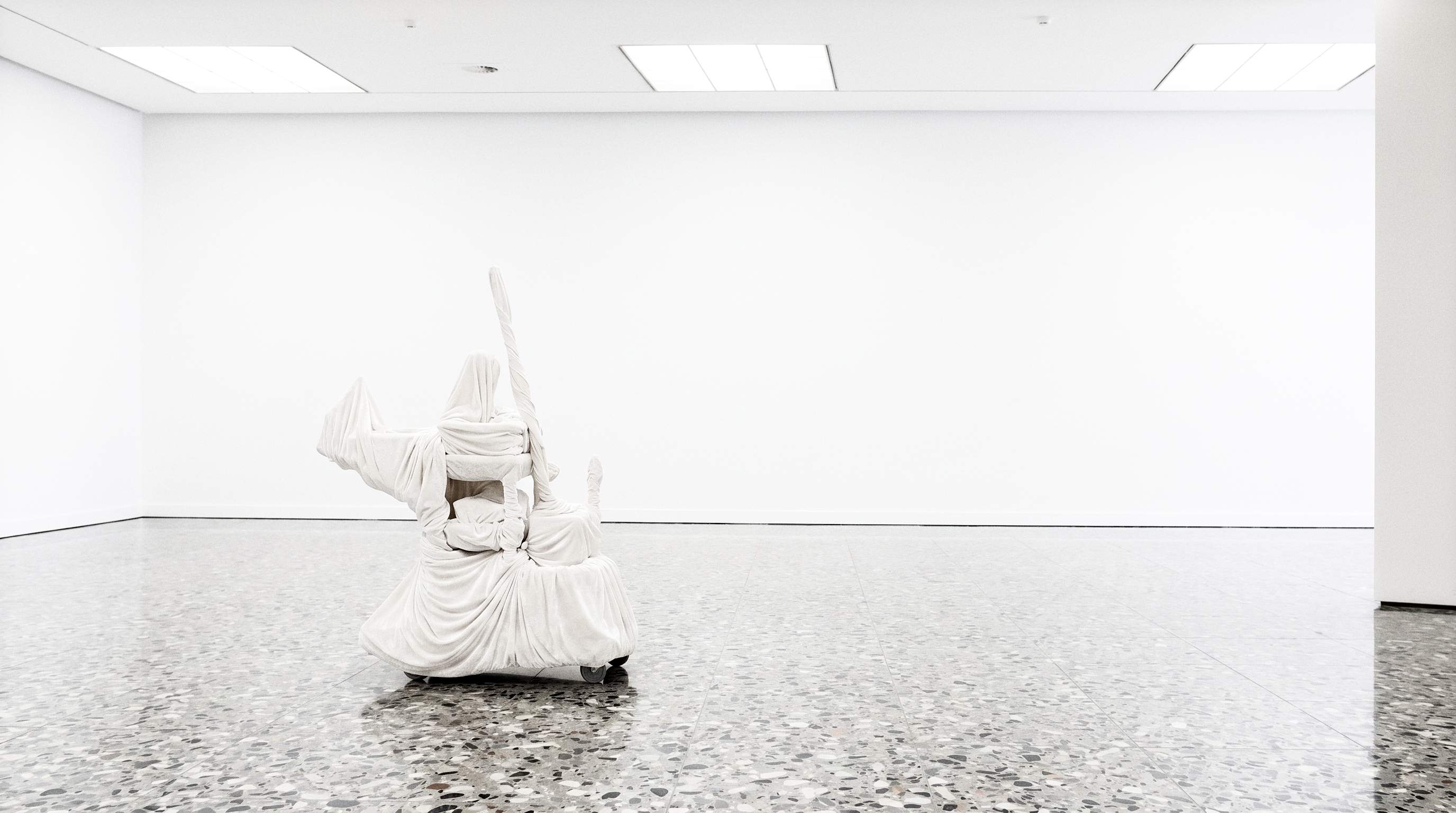 An empty white gallery with a terrazzo floor and a single white-shrouded form on castors standing mid-room.