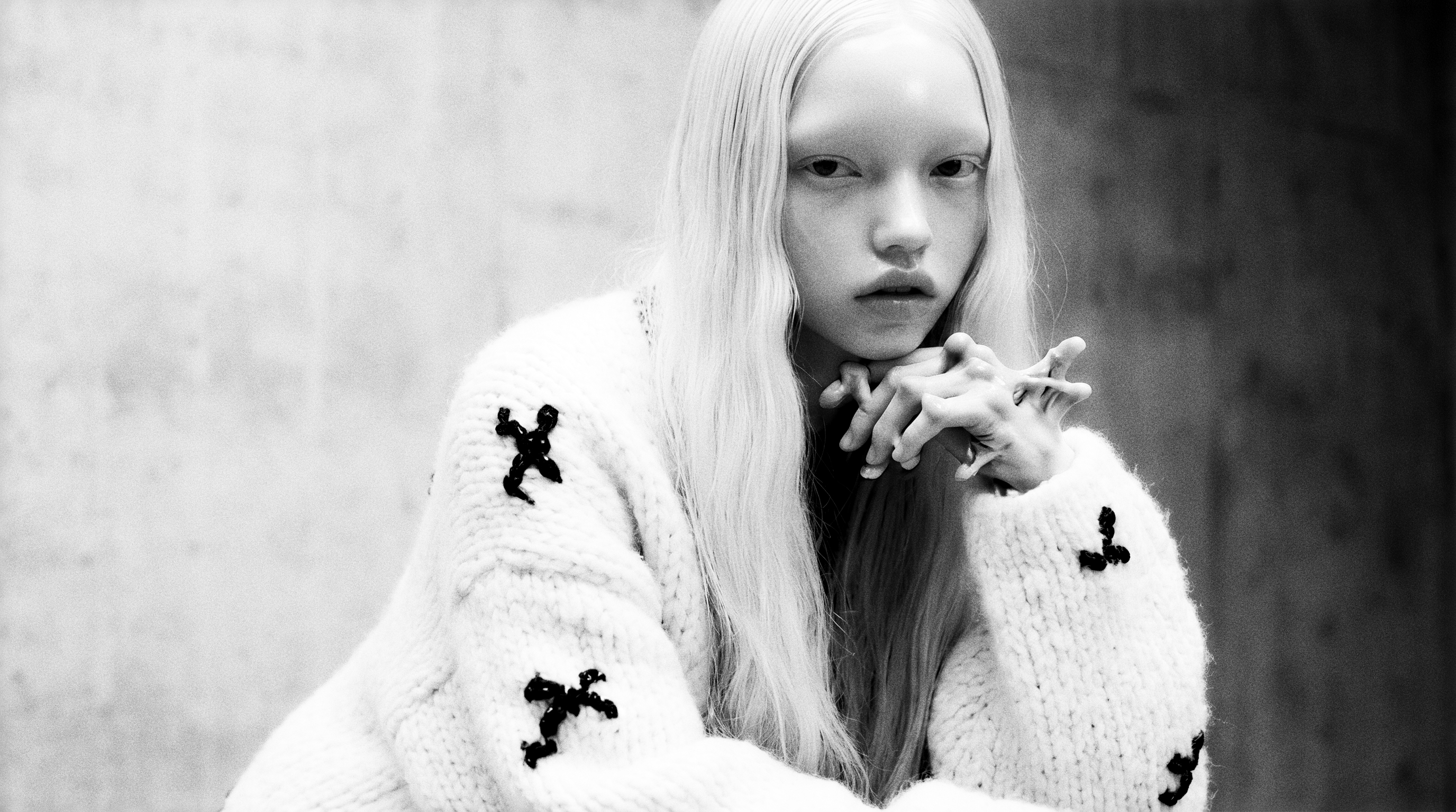 Black-and-white portrait of a pale platinum-haired figure in a chunky cream knit with black bows, chin resting on interlaced hands — the fingers tangling into one too many, the tell left in.