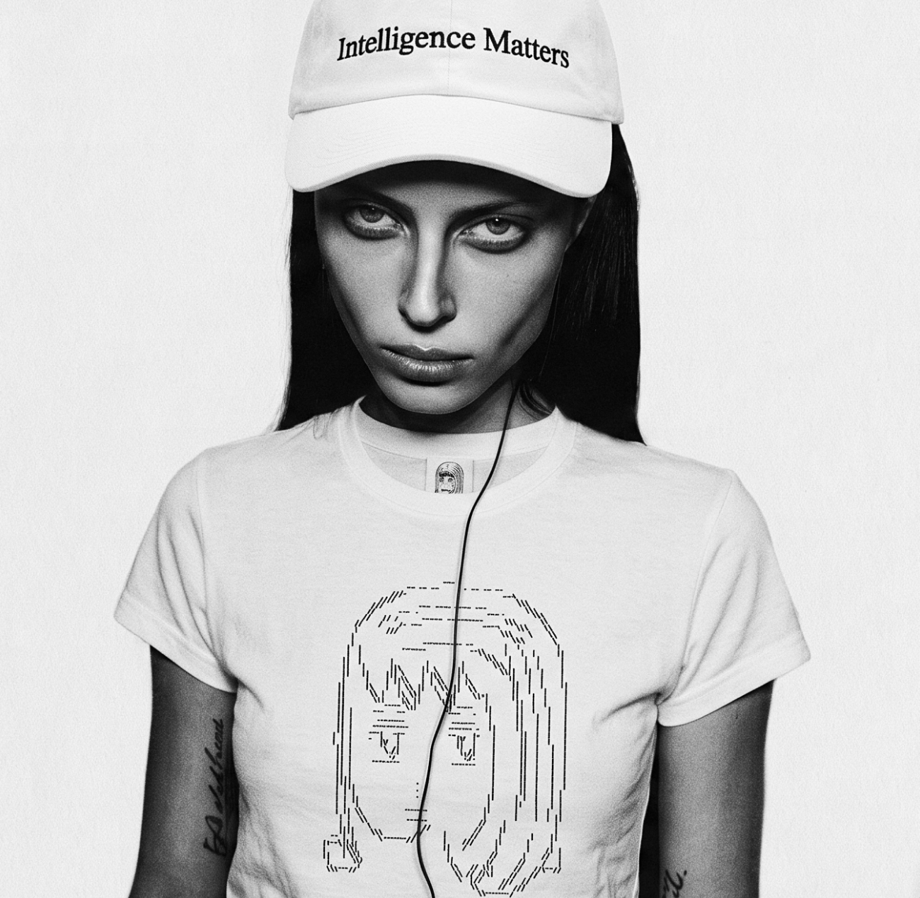 Black-and-white portrait of a young woman staring at the camera in a white Intelligence Matters cap and a white tee printed with an ASCII-rendered face; a small garment care-label sits on the front of her collar, at the throat.