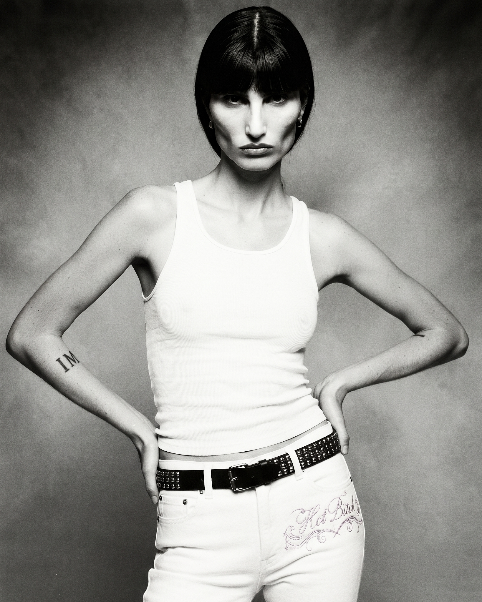 A monochrome woman with a blunt dark bob stands hands-on-hips in a white tank, low-rise jeans embroidered HOT BITCH in lavender and a black studded belt.