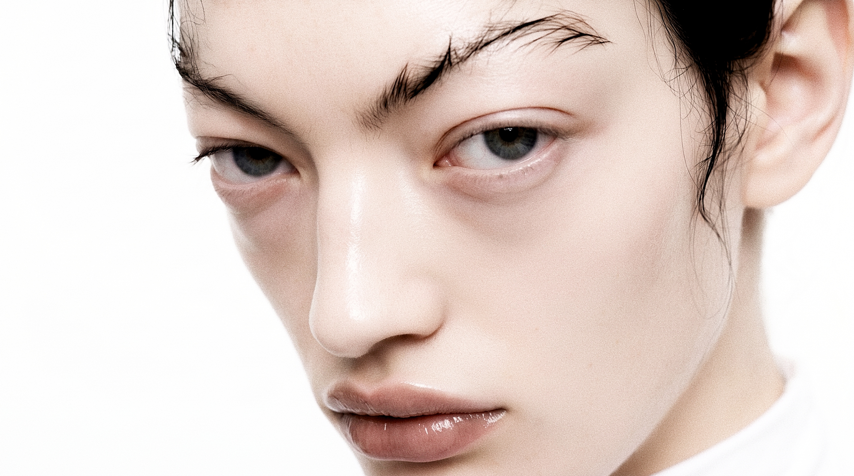 An extreme close portrait with wide grey-blue eyes, dark textured brows and glossy lips positioned at the right edge of a white field.
