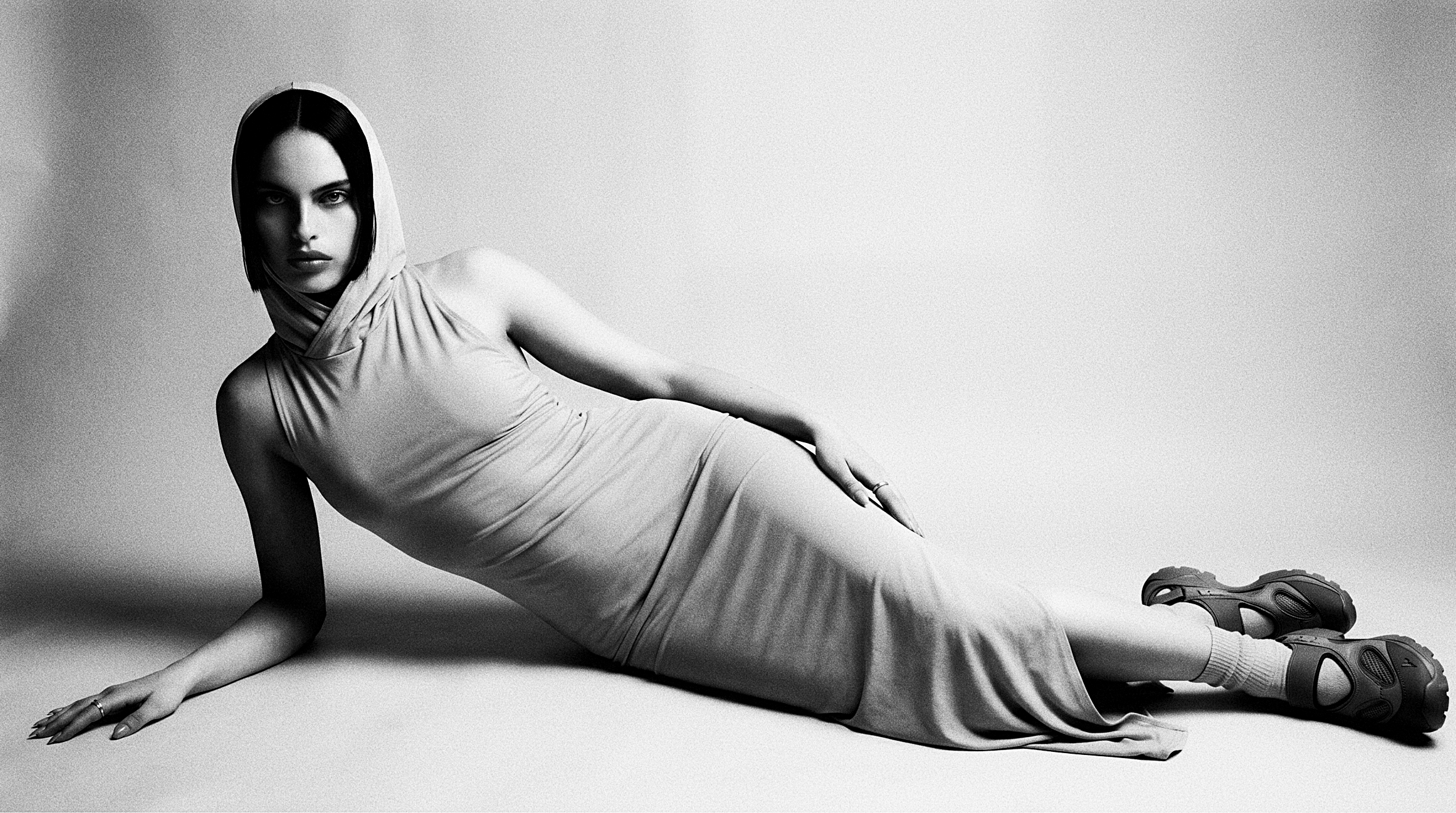 A hooded woman reclines diagonally on a white studio floor in a long pale dress, white socks and heavy hiking sandals.
