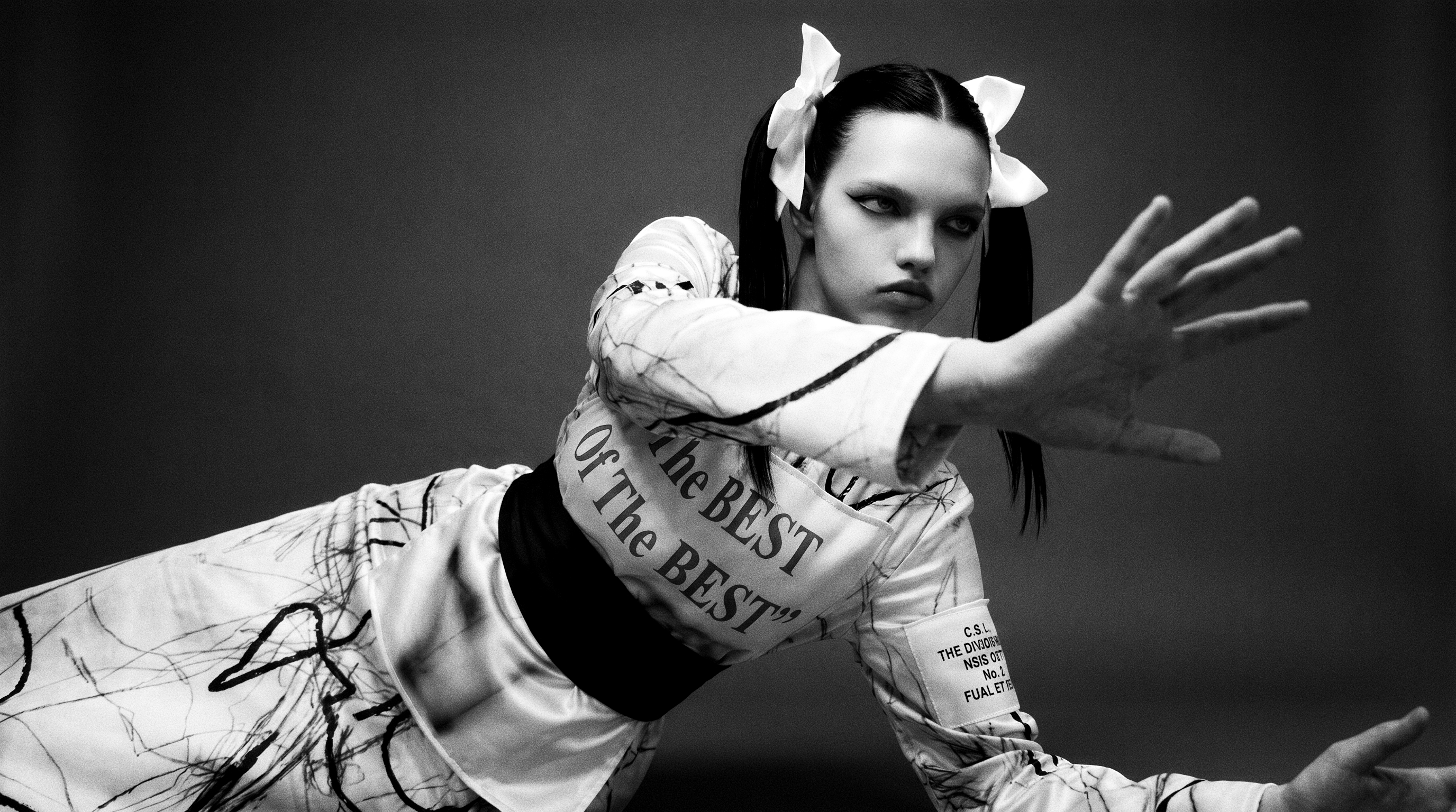 Black-and-white studio portrait — one figure with dark twin ponytails and large white bows reclines against a dark backdrop, reaching an open hand toward the lens in a white marked outfit printed ‘The BEST Of The BEST’ across the torso.