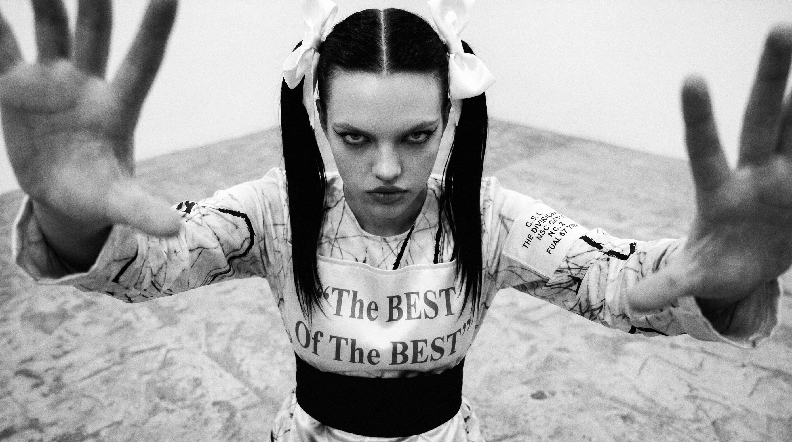 Black-and-white studio portrait — one figure with dark twin ponytails and large white bows faces the lens, extending both open hands toward the camera in a light patterned outfit printed ‘The BEST Of The BEST’ across the torso.