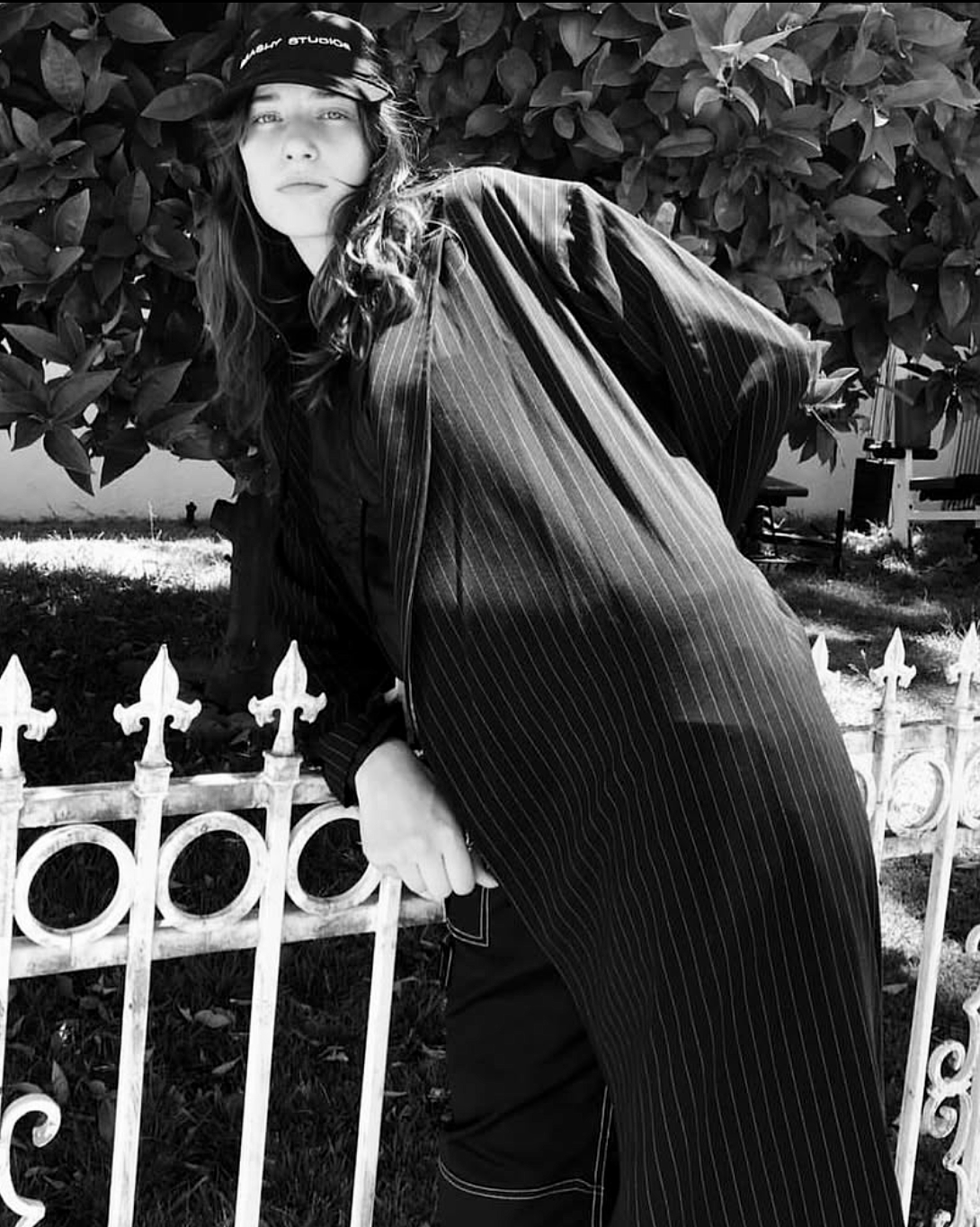 Black-and-white portrait in a Brashy Studios cap and oversized pinstriped shirt, leaning against a white iron fence.