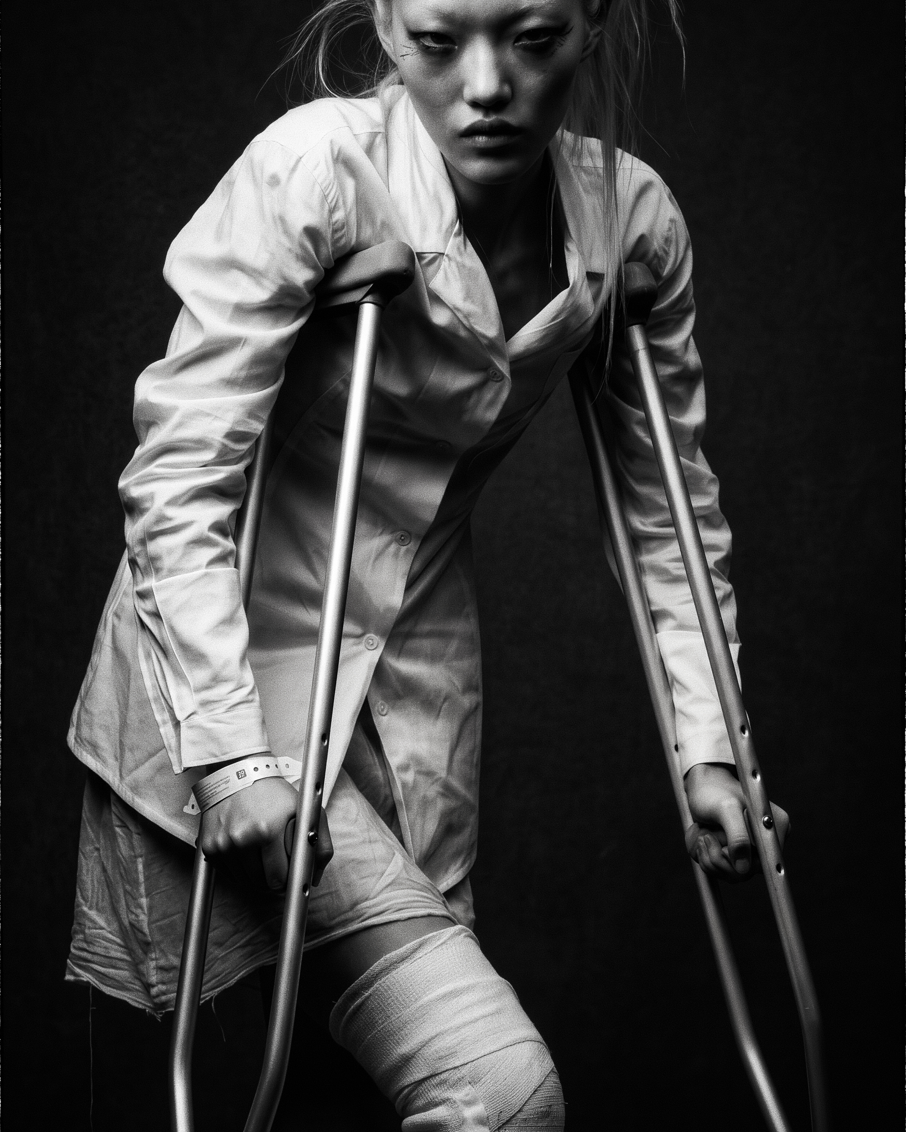 Black-and-white portrait of a figure on crutches, wearing a white shirt and a bandage around one knee.
