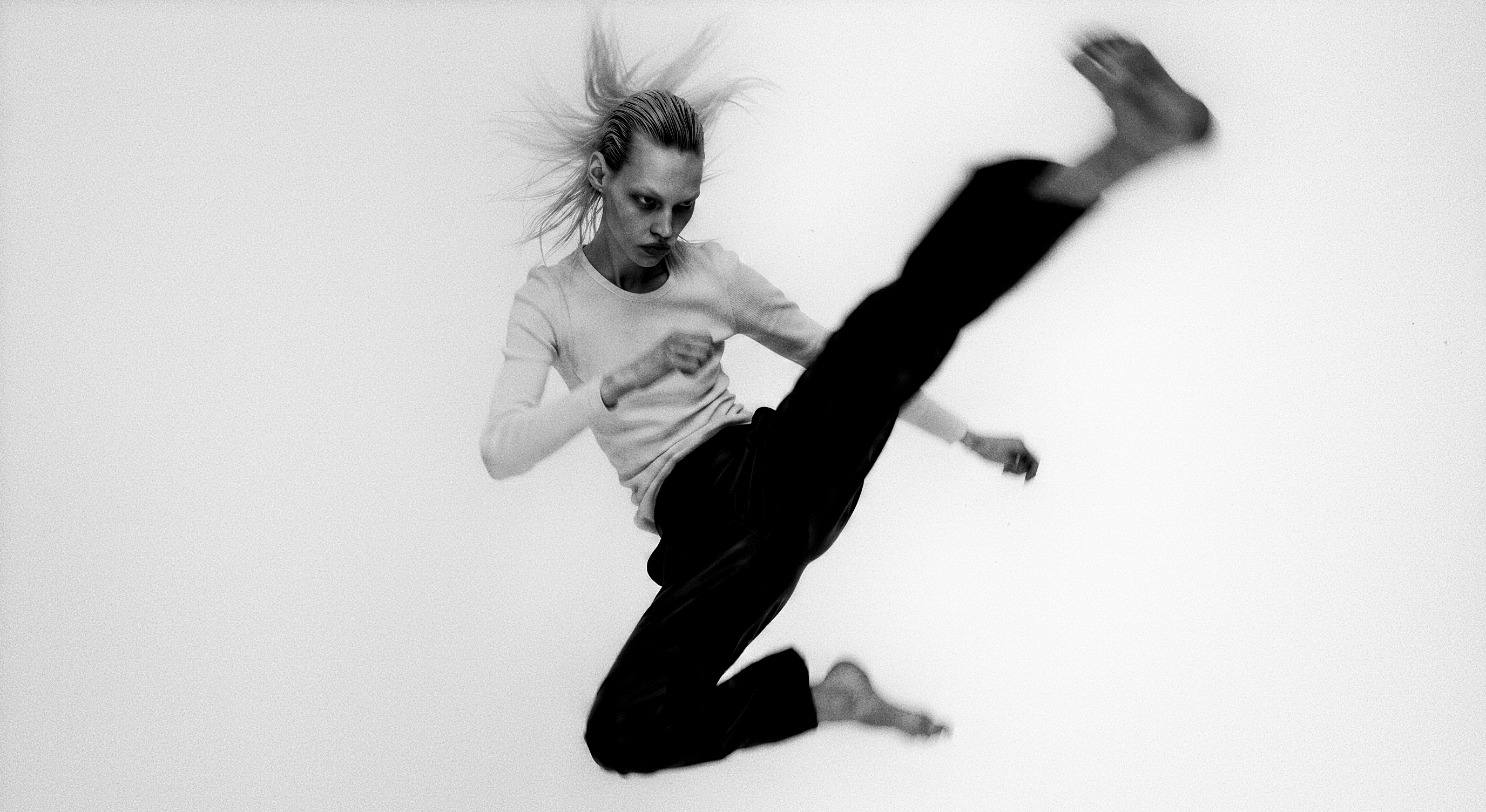 Person in a white long-sleeve top and black trousers kicking mid-air, hair blown back.