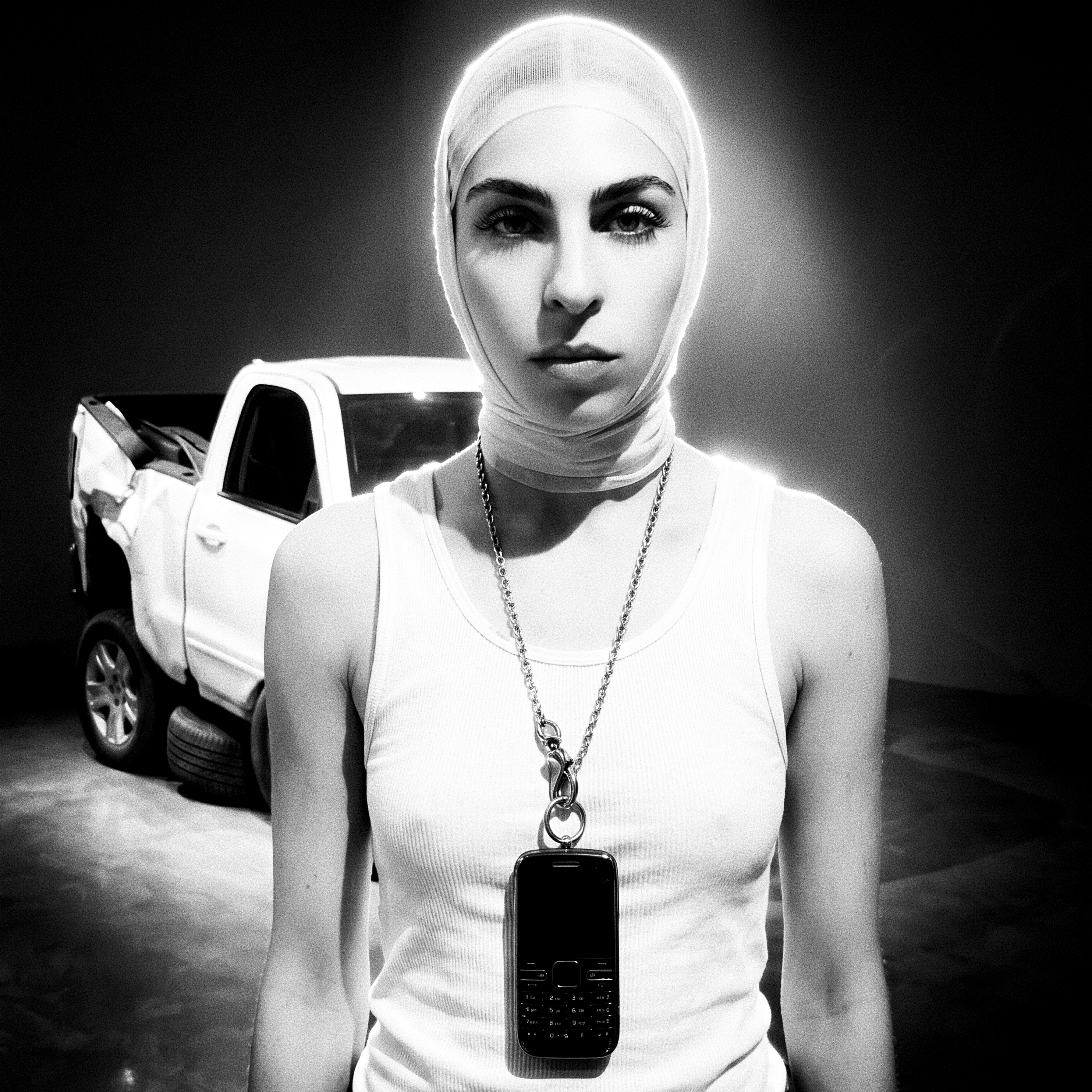 Leah McCarthy in a white mesh head-wrap and heavy silver chain suspending a keypad phone before a wrecked white pickup truck.