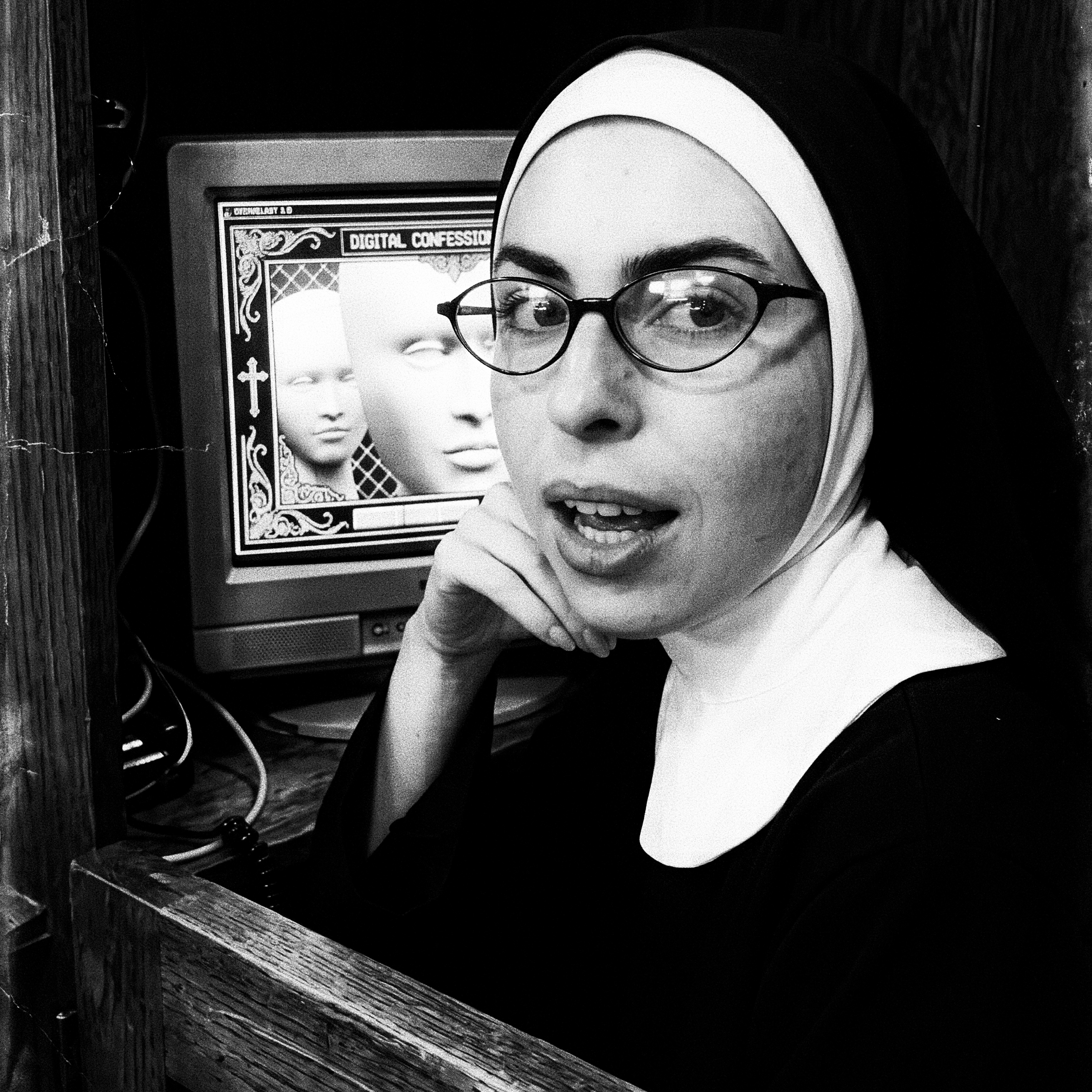 Leah McCarthy in a nun's habit and wire-rimmed glasses seated beside a CRT monitor running Cyberchurch 3.0 Digital Confession.