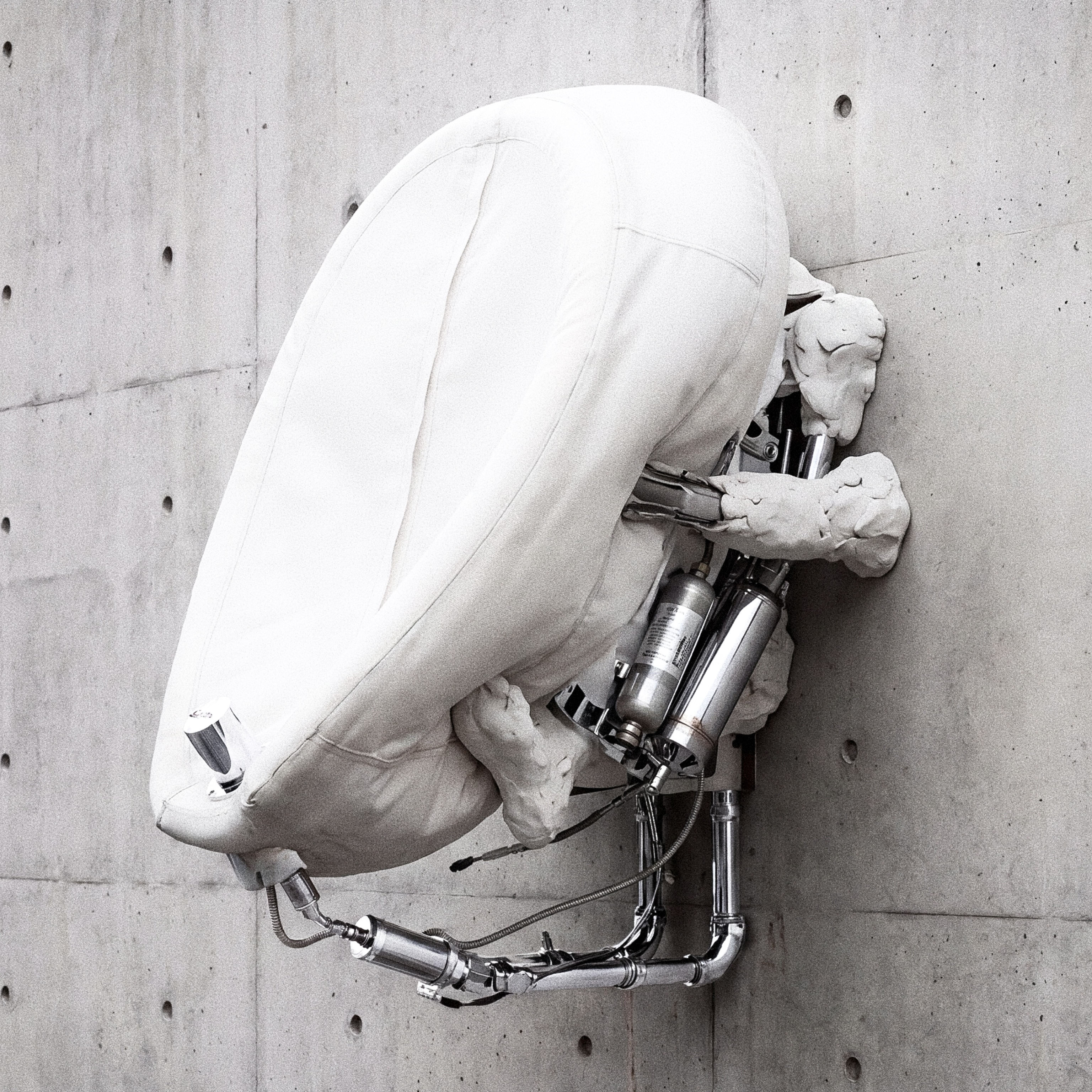 Wall-mounted white airbag cushion and plaster limbs pinned by stainless-steel pneumatic cylinders against board-formed concrete.