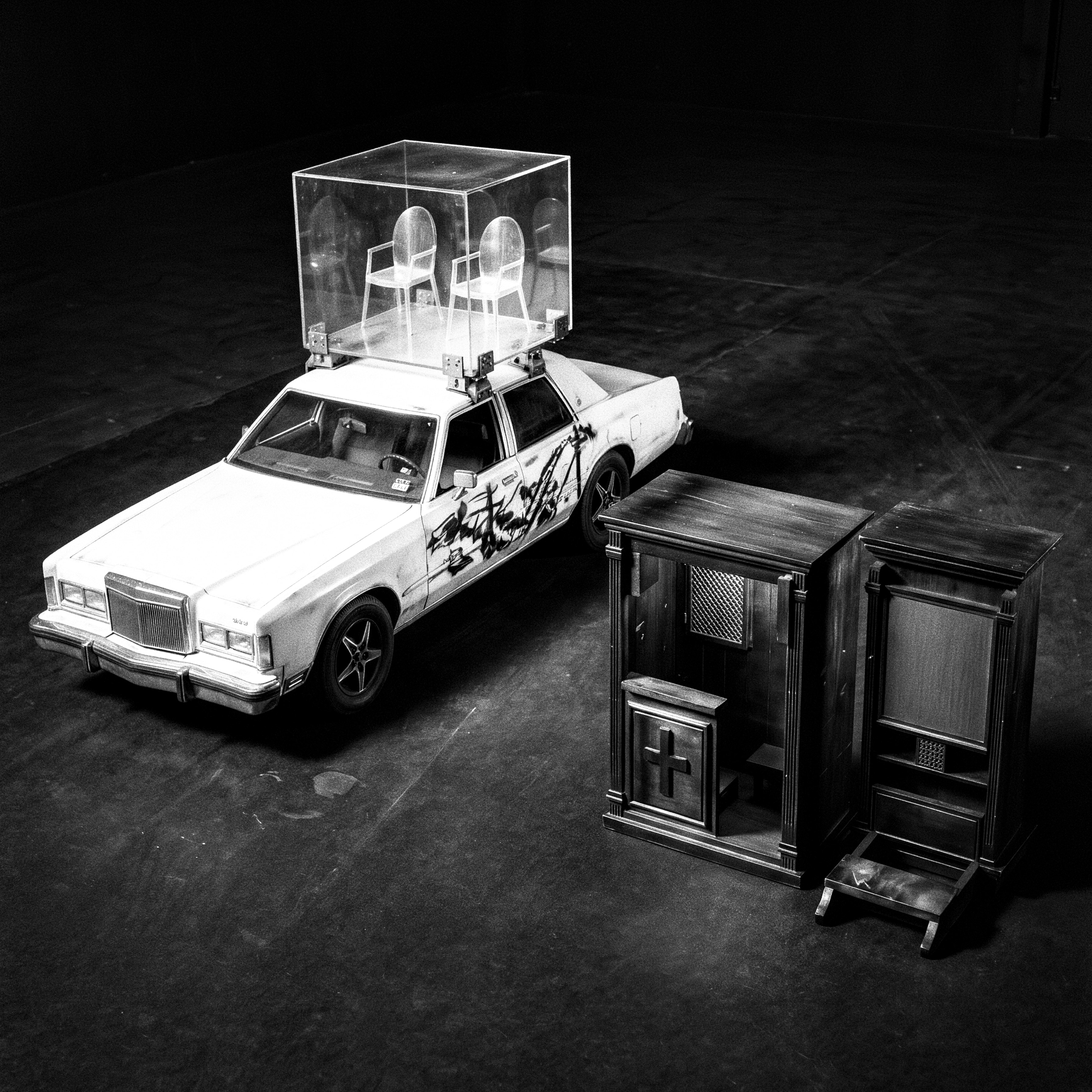 Confession Turk — white sedan carrying an acrylic roof vitrine of two transparent chairs beside two dark timber confession booths.
