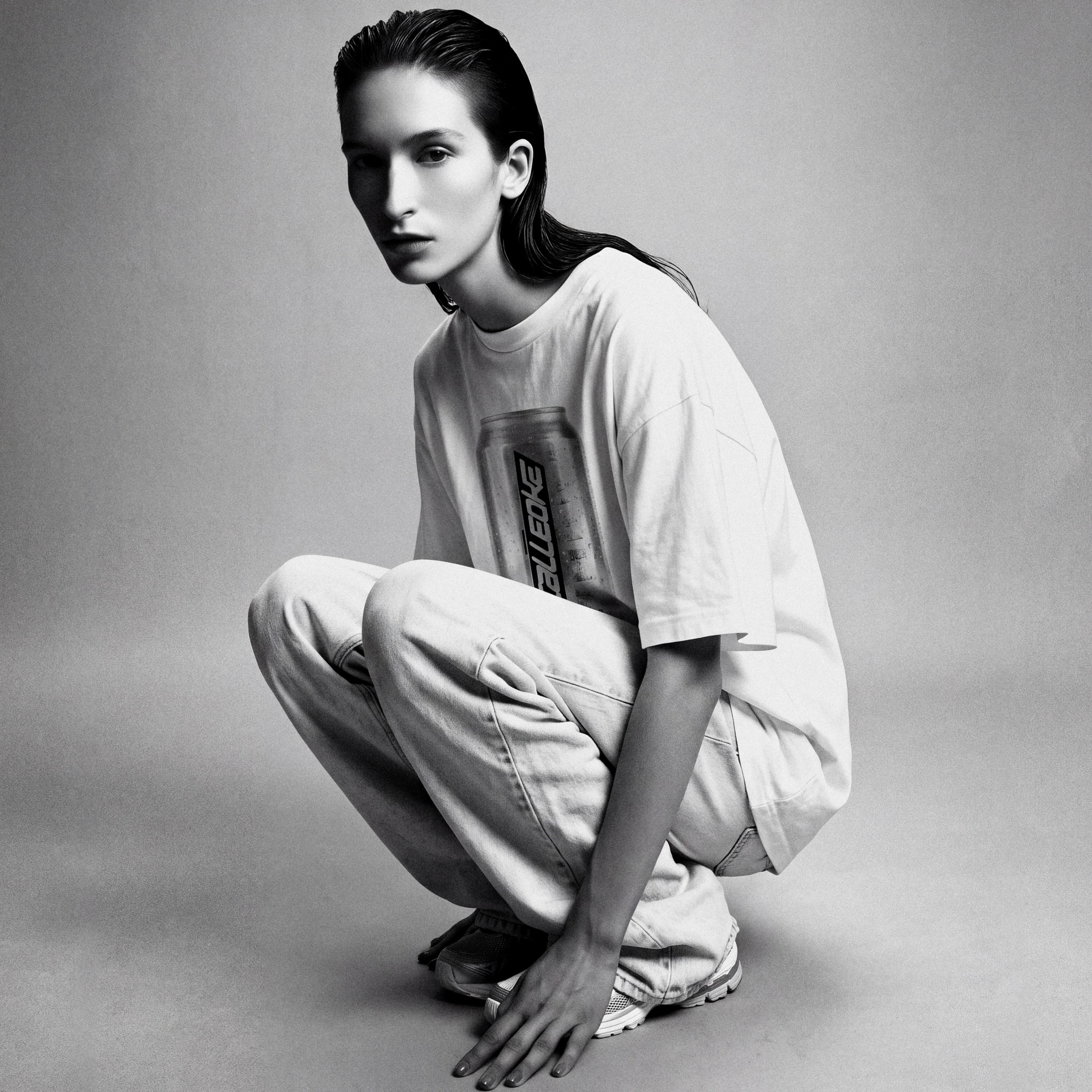 Dive Bar Plate 03 – monochrome studio portrait of a crouched figure in an oversized white cotton t-shirt printed with the Calleoke aluminium can, pale wide-leg denim and white trainers.