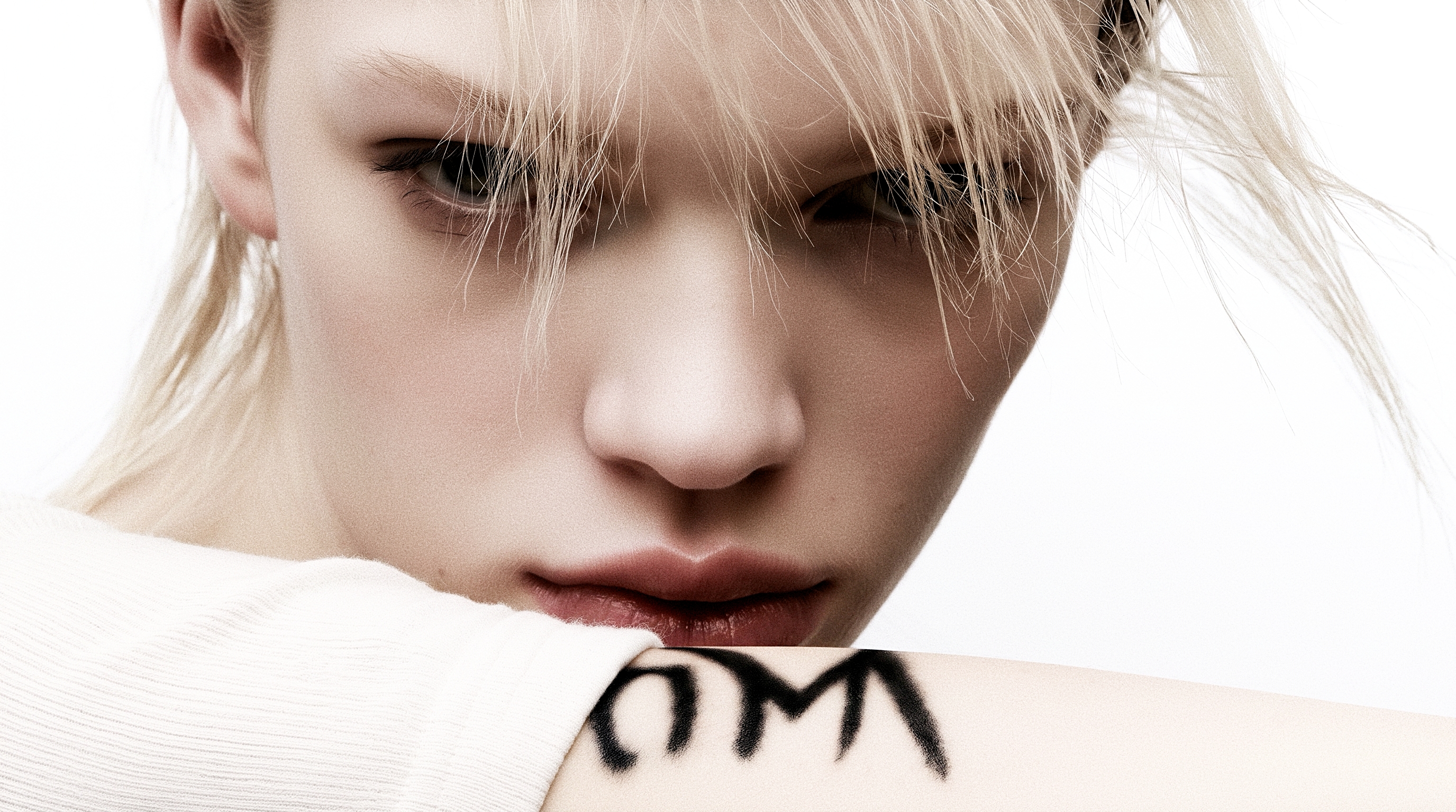 A close portrait of a platinum-haired woman in white ribbed knit with OM painted on her forearm and hair crossing one eye.